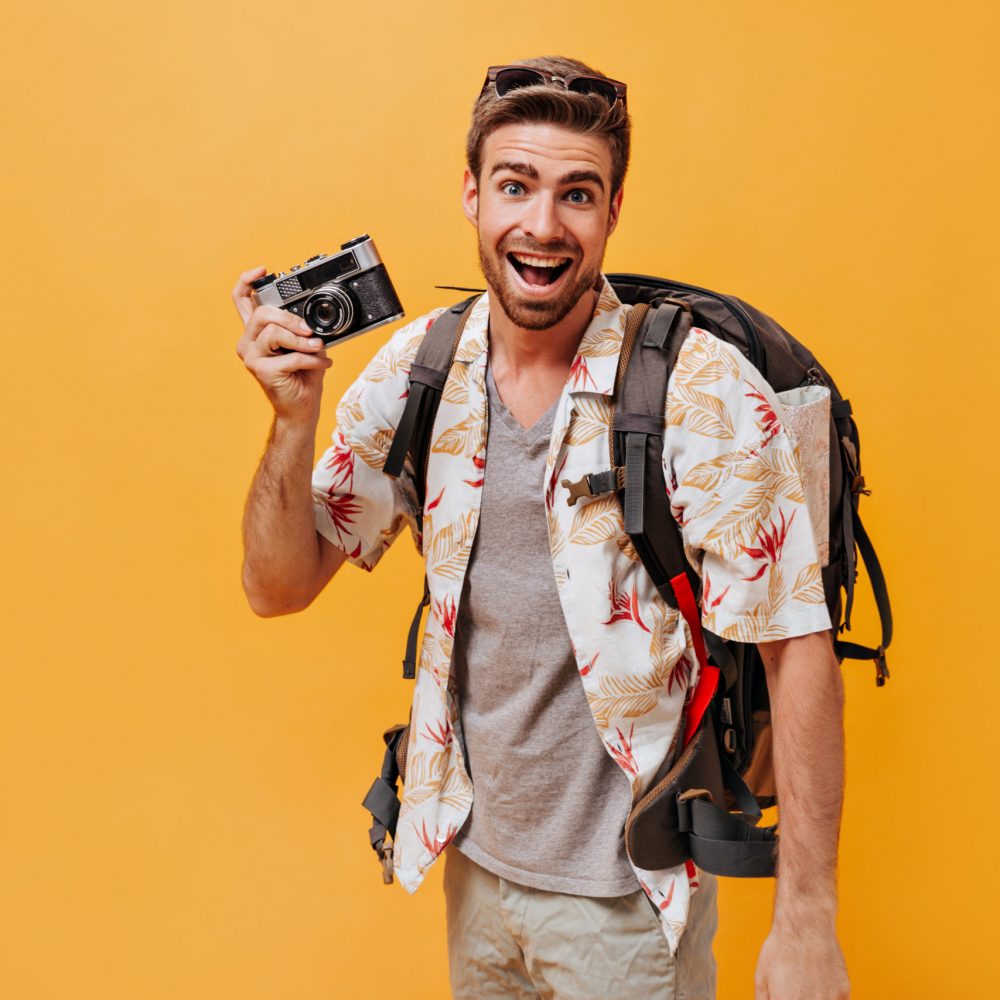 Cheerful man with beard in grey t-shirt and printed light shirt smiling and posing with camera and backpack on orange background..